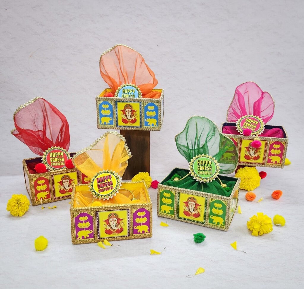 Floral Art presents A display of decorated gift boxes with colorful fabric coverings. Each box features an image of Lord Ganesha and a badge labeled "Happy Ganesh Chaturthi." Yellow and orange flowers are scattered around.