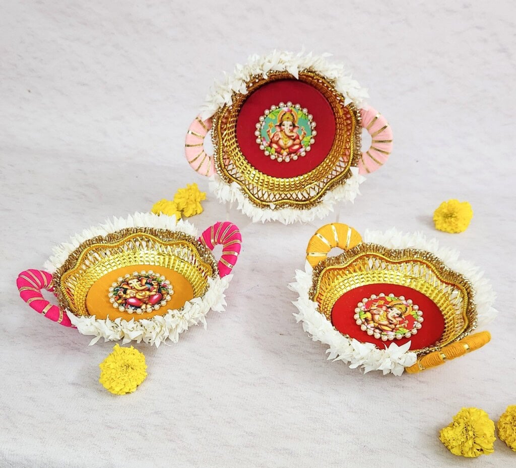 Floral Art presents Three decorative plates with intricate beadwork, white flower borders, and deity images are displayed on a light colored surface, accompanied by scattered yellow flowers.