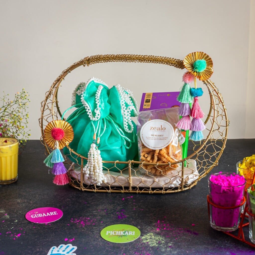Floral Art presents A decorative basket with a teal bag, a packet of snacks, and colorful tassels. It is surrounded by bright powders and drinks, suggesting a festive or cultural celebration.