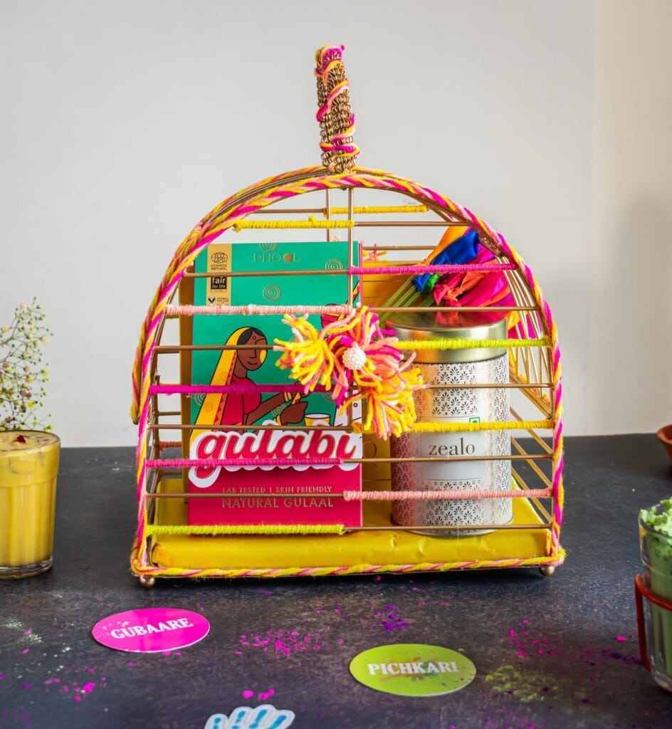 Floral Art presents A colorful gift basket with Holi themed items including boxes, a jar, and decorative elements. Coasters with "Gulabi" and "Pichkari" text are on the table.