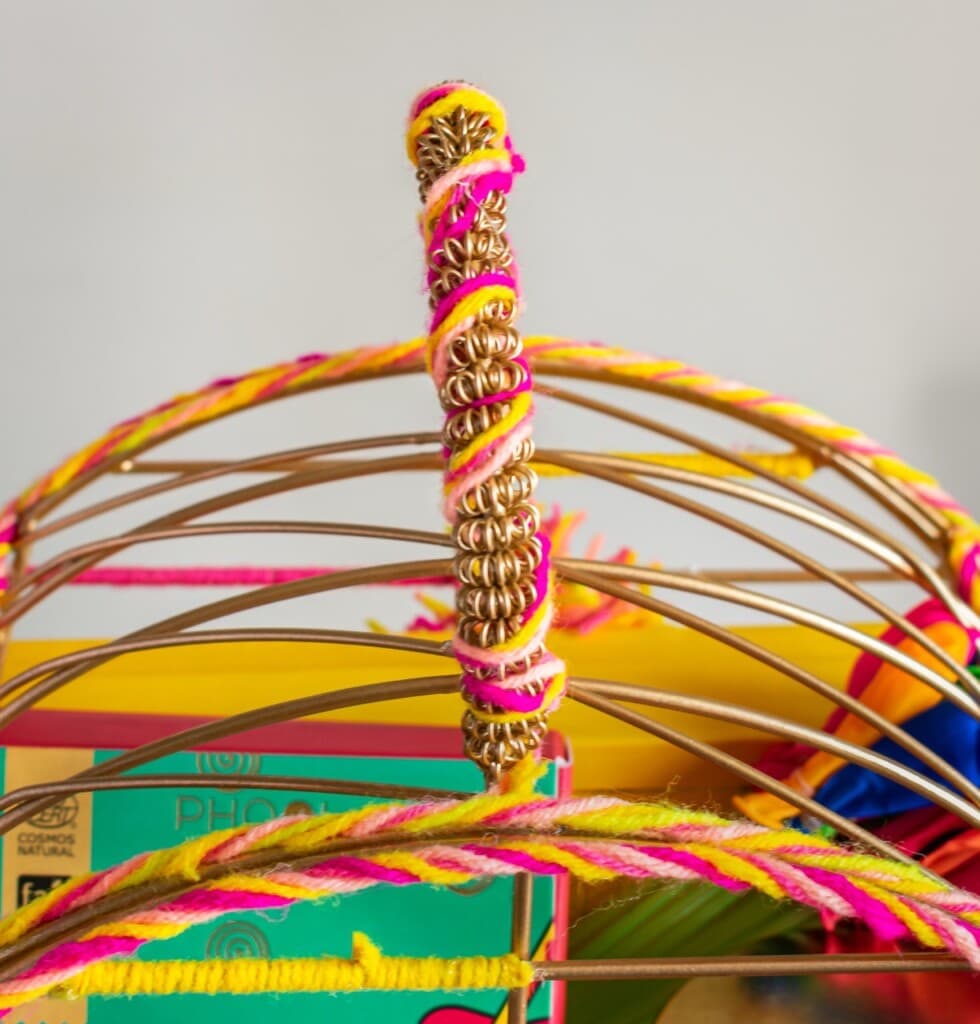 Floral Art presents Woven metal handle of a birdcage wrapped in pink and yellow yarn, with colorful objects inside.