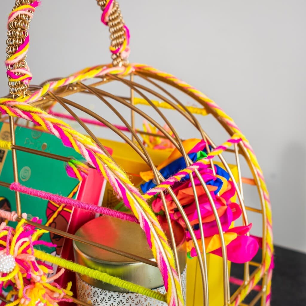 Floral Art presents A colorful, decorated cage filled with assorted items, including balloons, a tin, and a book, placed against a plain background.