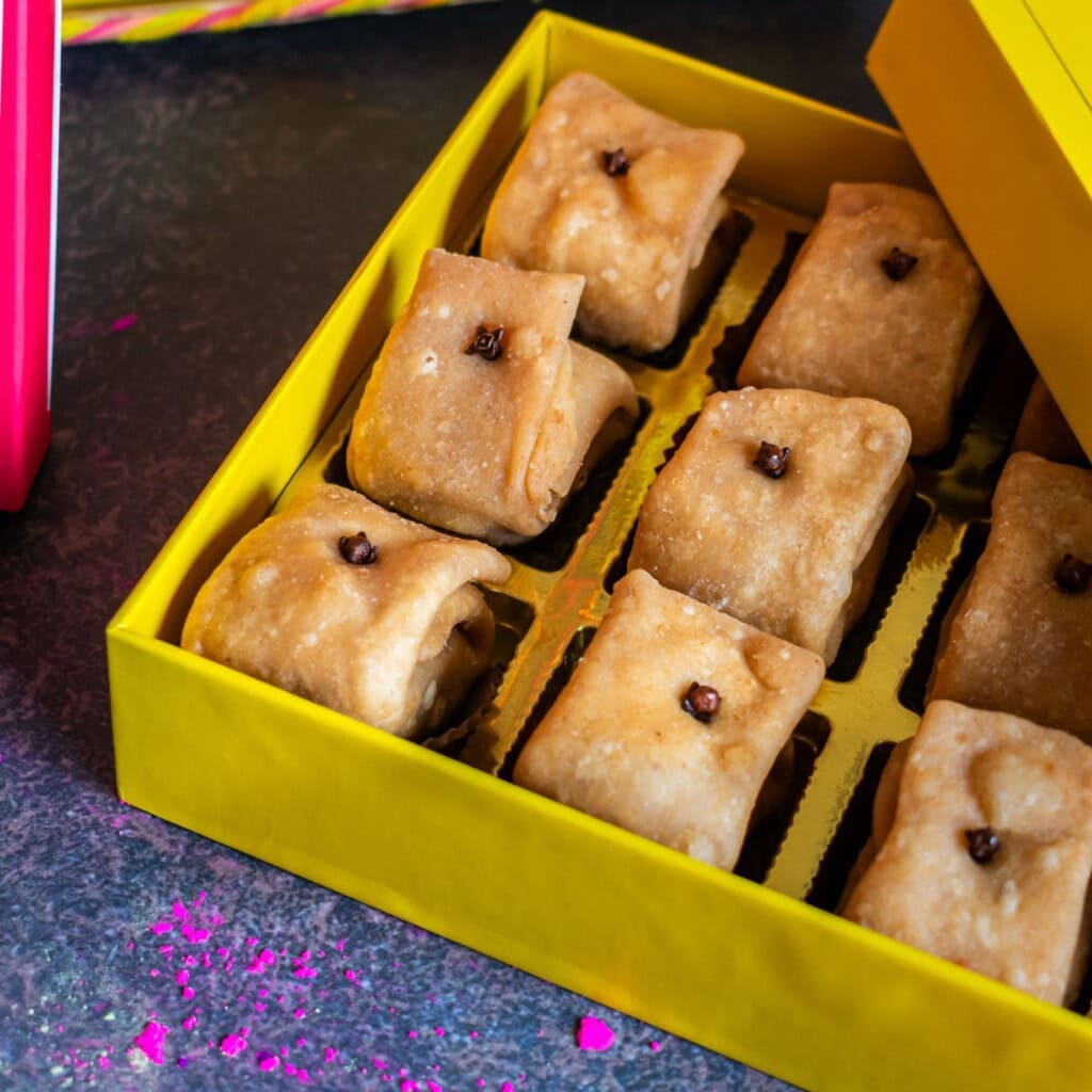Floral Art presents A yellow box containing neatly arranged pieces of Indian sweets stuffed and folded, with cloves on top, placed on a dark surface.
