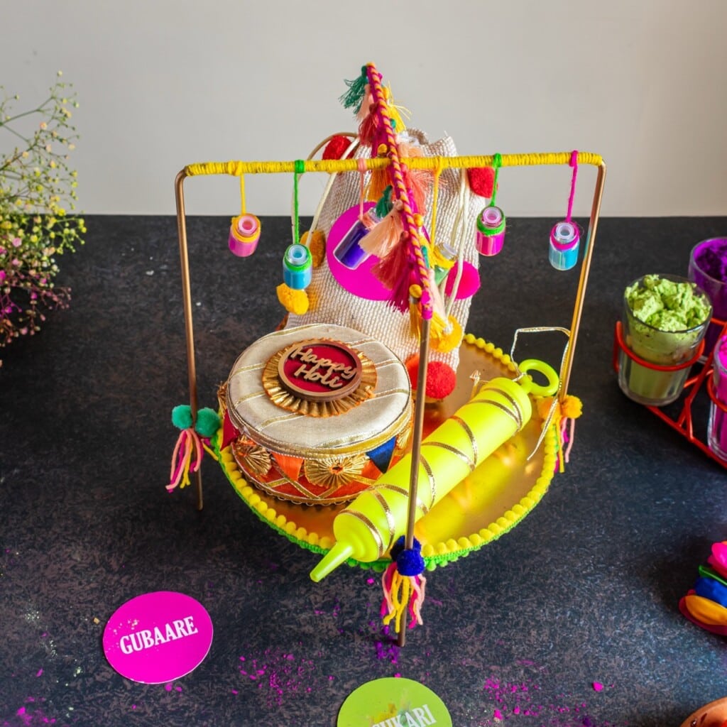 Floral Art presents Colorful Holi themed decor including a dhol, pichkari (water gun), and decorative elements displayed on a black surface.