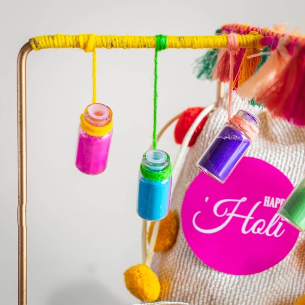 Floral Art presents Three small jars filled with pink, blue, and purple powder hang from a yellow yarn on a frame. A sack with "Happy Holi" printed on it is in the background.