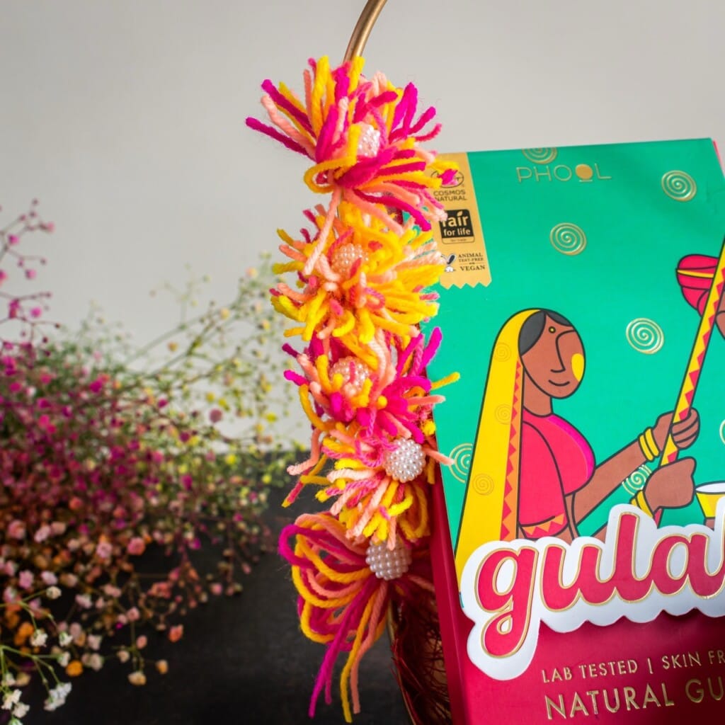 Floral Art presents Colorful yarn decoration and a box with vibrant artwork featuring a person in a sari, labeled "natural gulal." Background has soft focus flowers.