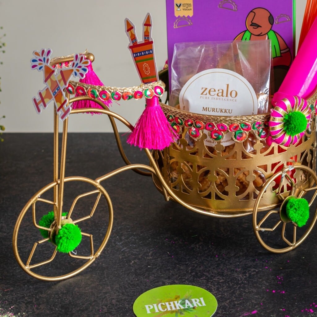 Floral Art presents A decorative golden tricycle holds a pack of Murukku and pink party items, adorned with colorful tassels and embellishments.
