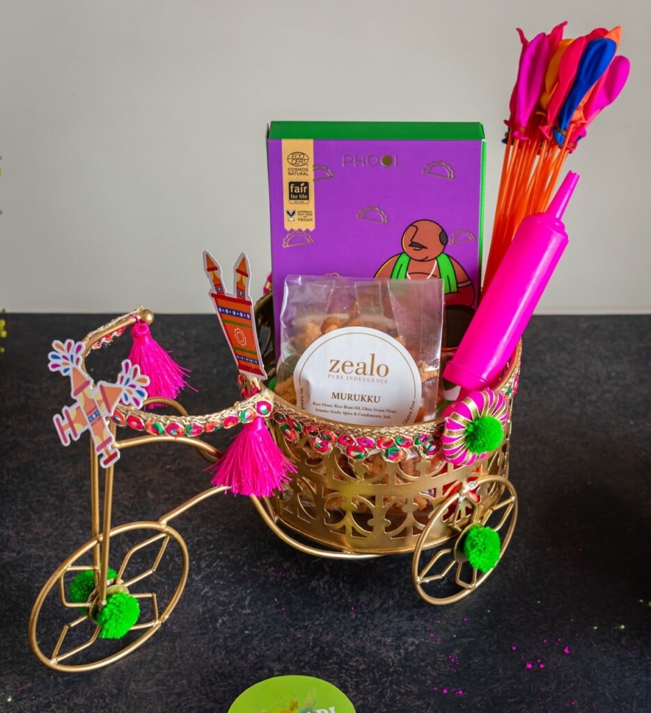 Floral Art presents Decorative tricycle holds a gift basket with colorful items, including a box, Murukku snack, a toy rolling pin, and vibrant balloons, set against a neutral background.
