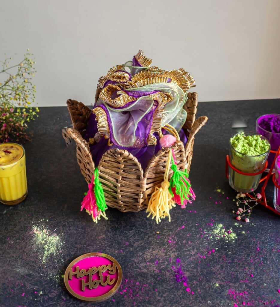Floral Art presents Wicker basket with colorful cloth, surrounded by Holi powders, a "Happy Holi" sign, and a yellow drink on a dark surface.