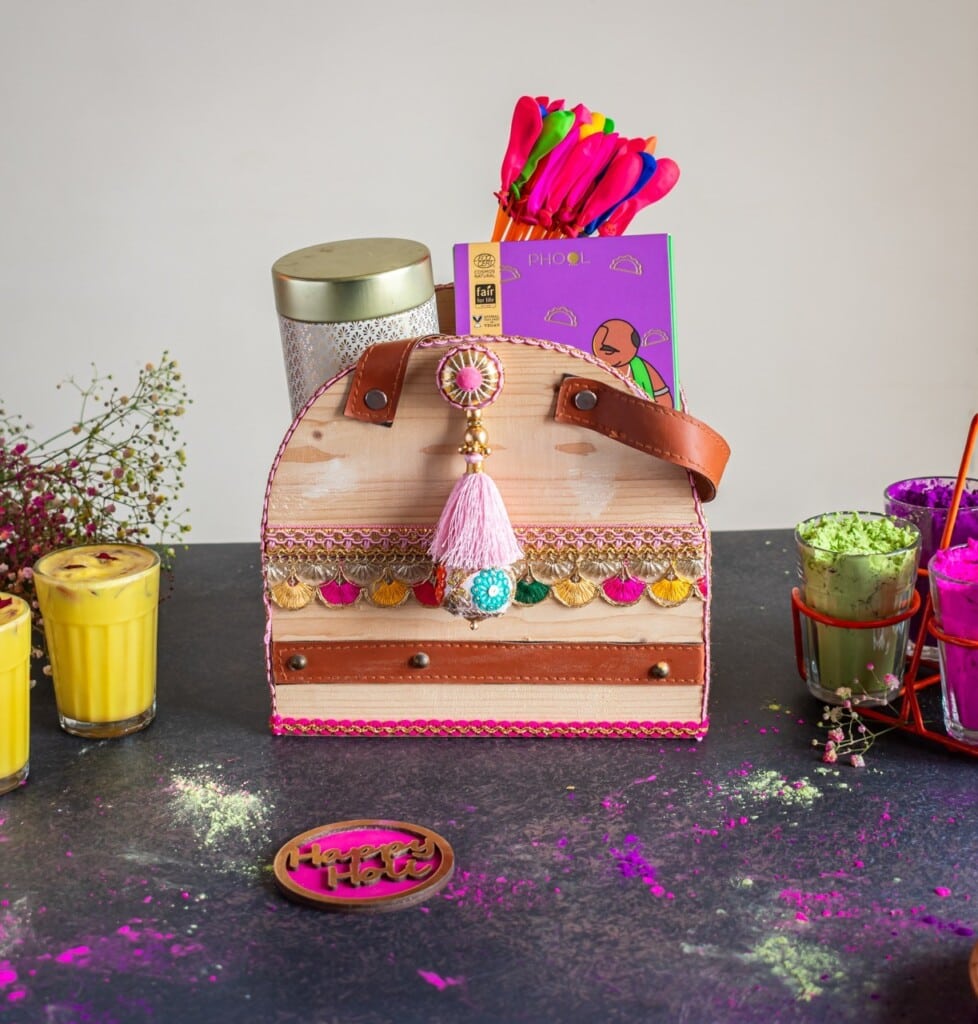 Floral Art presents A decorative bag with a tassel is surrounded by colorful drinks, a metal container, and bowls of colored powders on a table. A card and colorful sticks are inside the bag.