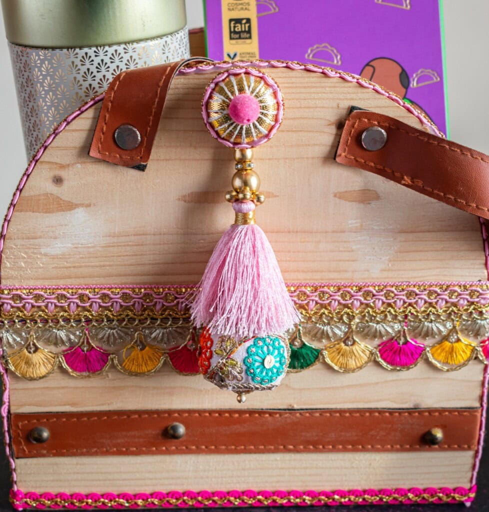 Floral Art presents Decorative wooden container with pink tassel and colorful accents, featuring leather straps and floral patterns, placed in front of a purple and white patterned background.