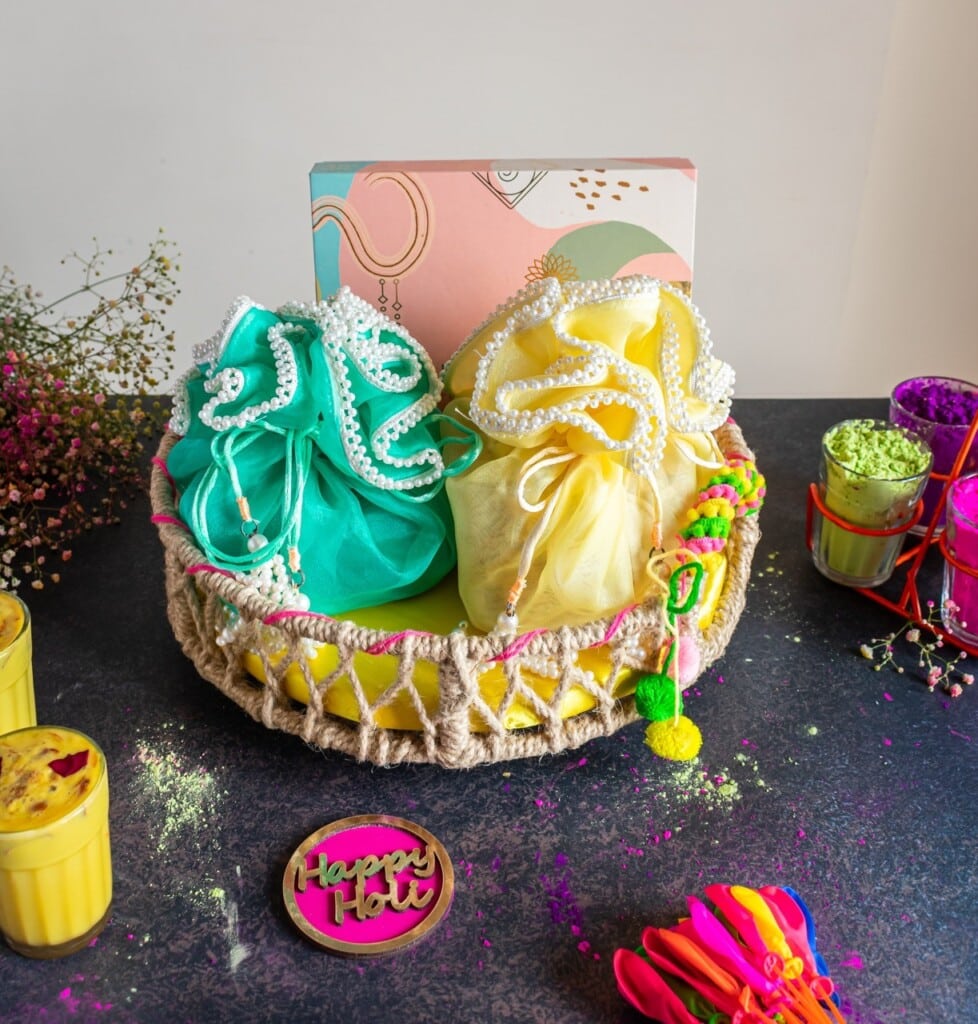 Floral Art presents A colorful basket with two wrapped gifts, surrounded by Holi powders, drinks, flowers, and a "Happy Holi" sign on a black surface.