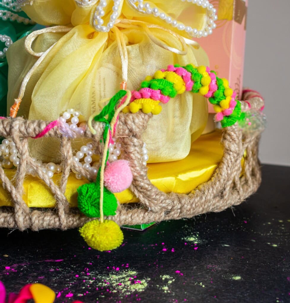 Floral Art presents A decorative basket with colorful pom poms and a yellow gift bag inside, set against a dark surface with scattered powder.
