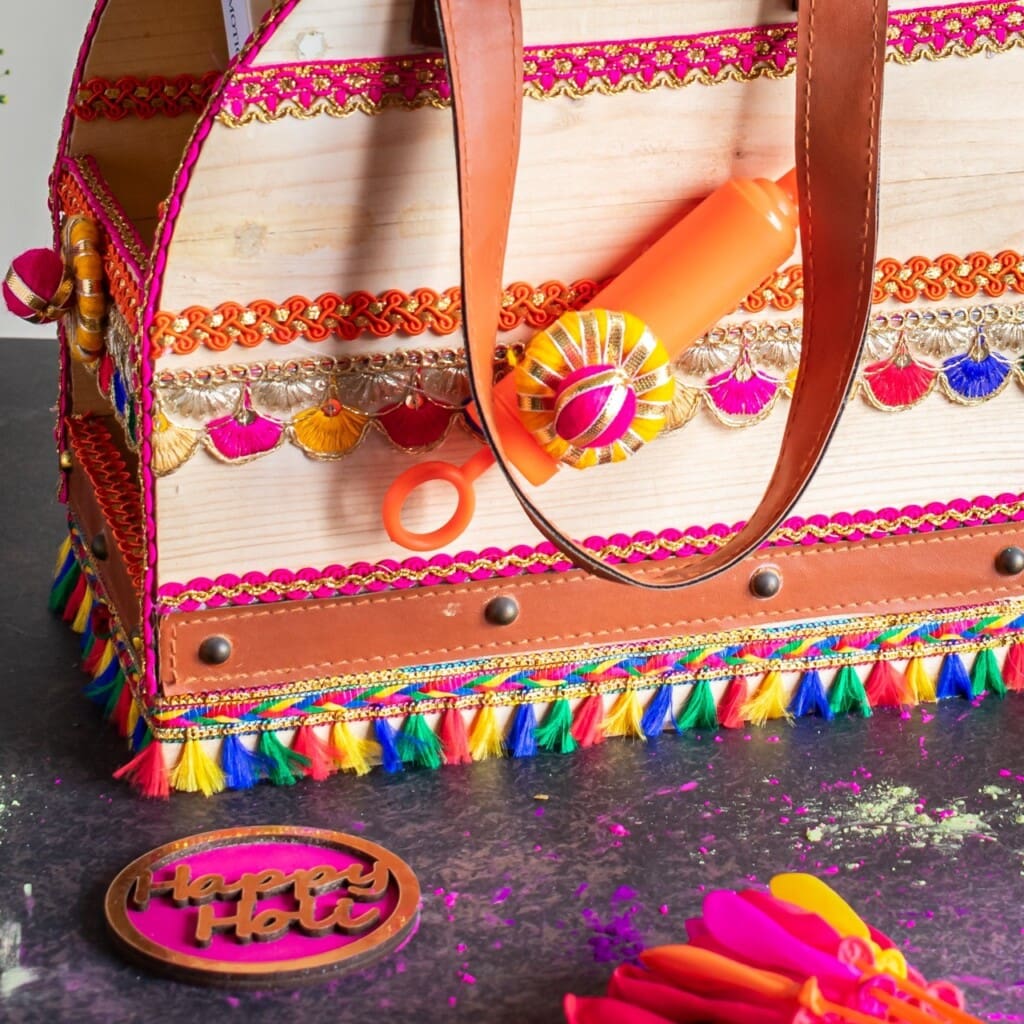 Floral Art presents Decorative box like container adorned with multicolored tassels, ribbon, and sequins. It holds a vibrant water spray bottle. Nearby, a "Happy Holi" coaster and colorful powders.