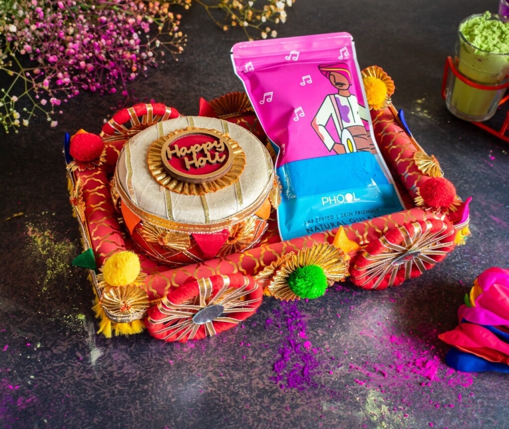 Floral Art presents Festive Holi gift basket with decorated container, pink gift bag, and colorful powders scattered on a dark surface.