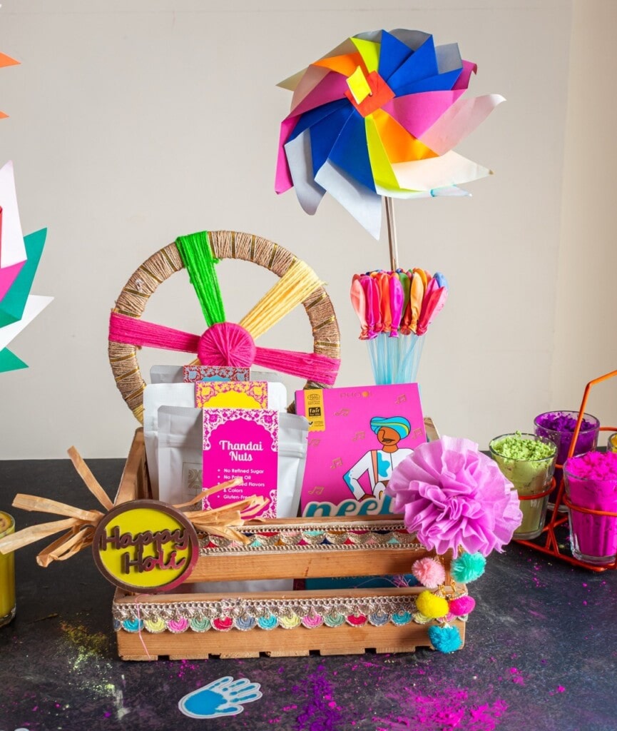 Floral Art presents A colorful Holi themed gift basket with snacks, colorful powders, and a paper pinwheel. Decorated with pom poms and a "Happy Holi" sign.