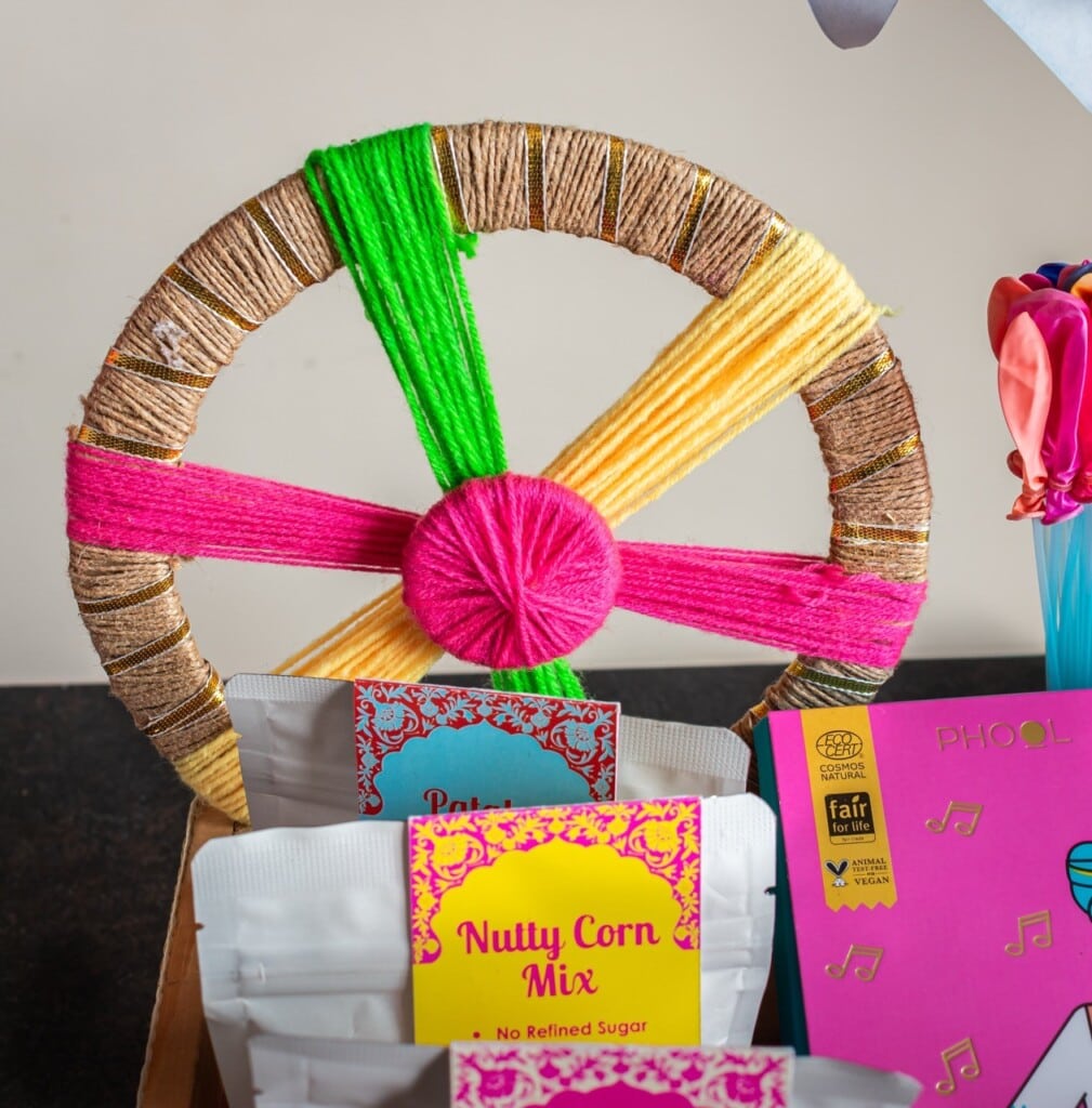 Floral Art presents Colorful yarn wrapped wheel with snack packets displayed in front, including "Nutty Corn Mix" and "Phool" packaging.
