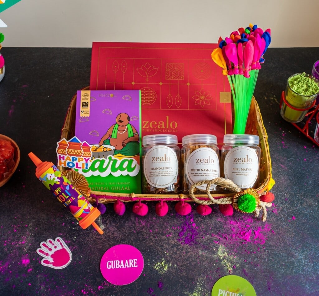 Floral Art presents A festive gift basket with colorful boxes, jars, a "Happy Holi" toy, and colorful props on a dark surface with scattered colored powders.