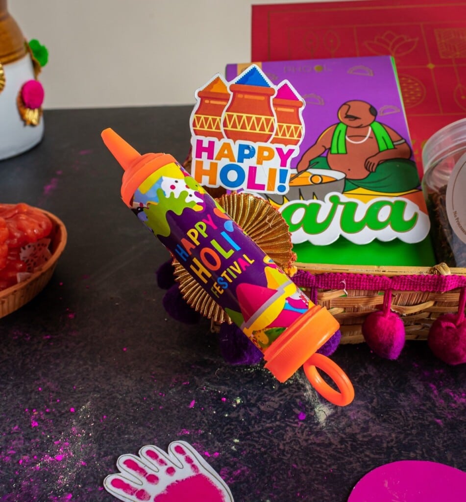 Floral Art presents Colorful "Happy Holi" pichkari surrounded by festive items, including a basket, pom poms, and packets on a table.
