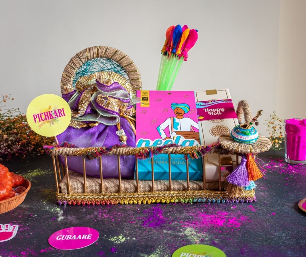 Floral Art presents A festive basket with colored powders, snacks, and water balloons, adorned with tassels and colorful decorations. Signs say "Pichkari," "Gubaare," and "Neela.