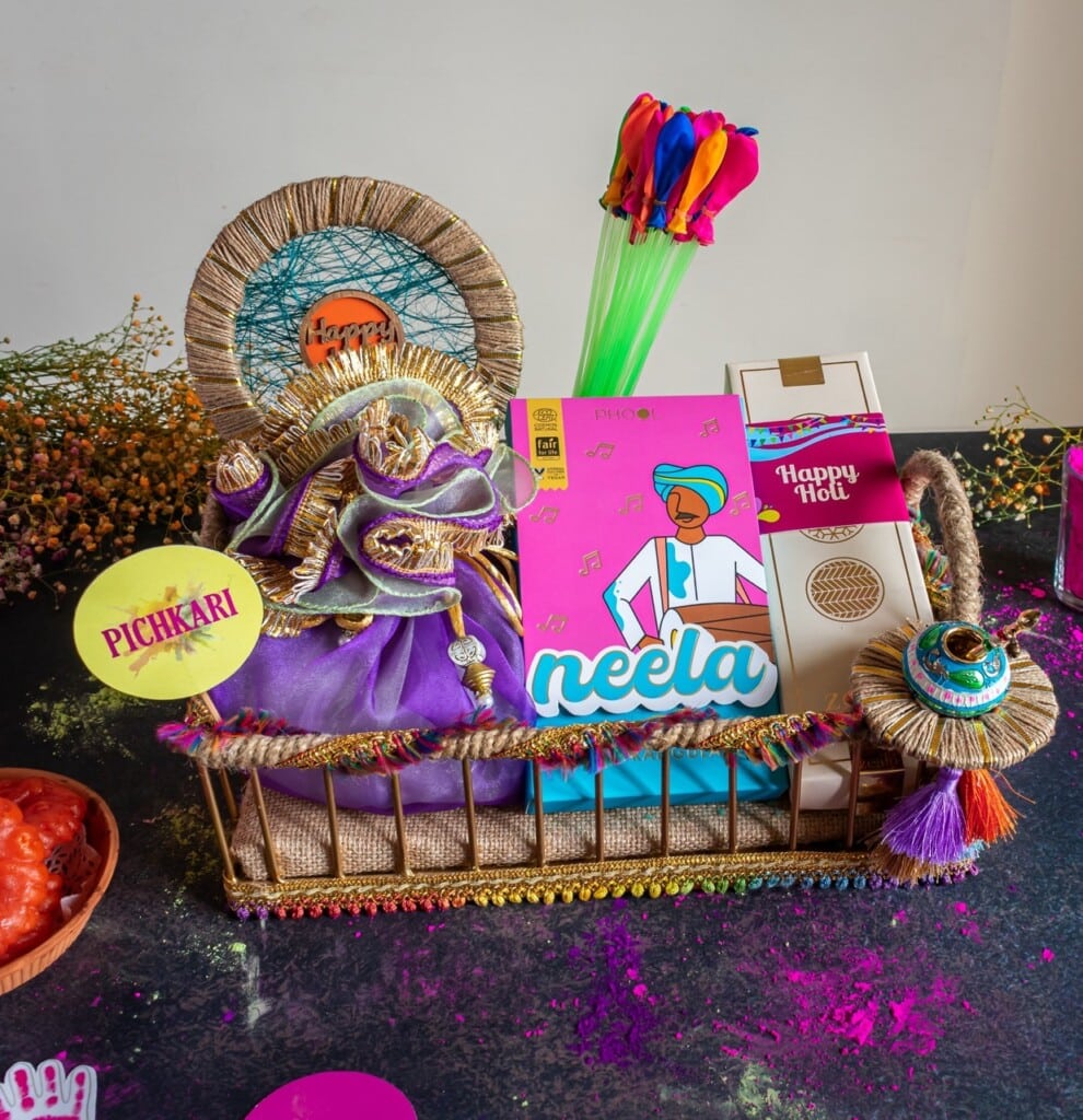 Floral Art presents A basket with Holi items: colorful balloons, snack boxes, a purple pouch, a "Happy Holi" tag, and decorative elements on a dark surface.
