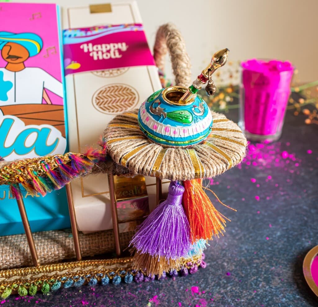 Floral Art presents A colorful Holi themed gift display featuring a decorative matka, vibrant tassels, a box of snacks, and a glass filled with pink powder on a dark surface.