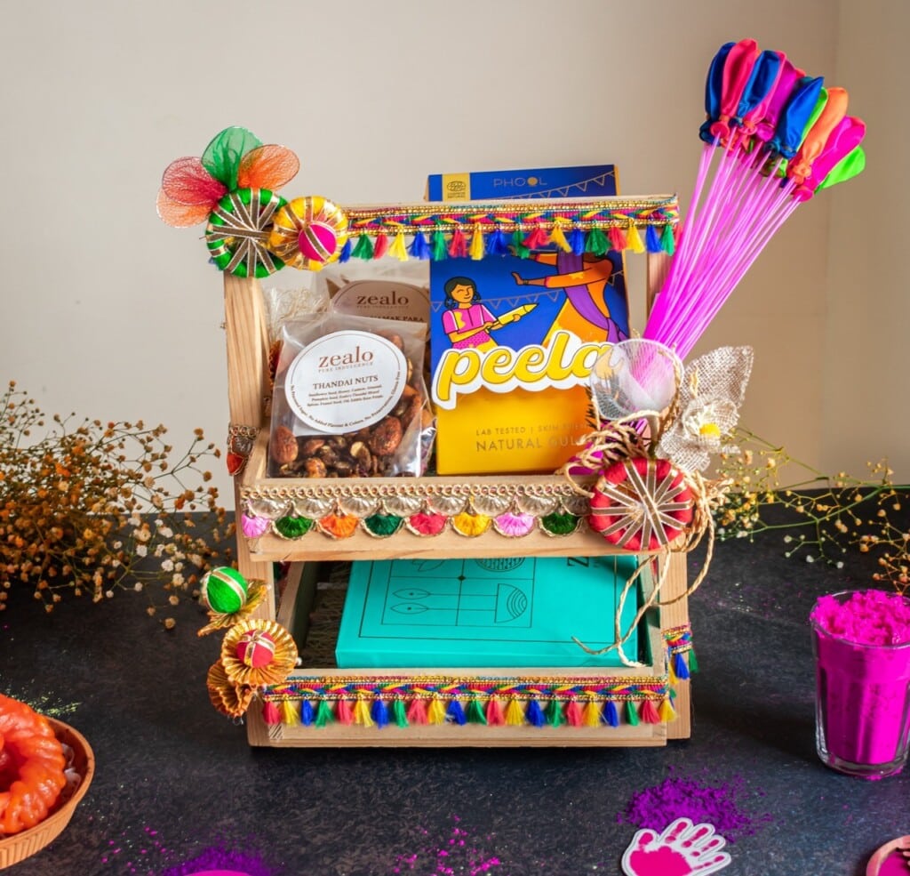 Floral Art presents Wooden shelf adorned with colorful decorations, holding snacks, a drawing book, and balloons. Nearby are vibrant powders and sweets, suggesting festive celebration.