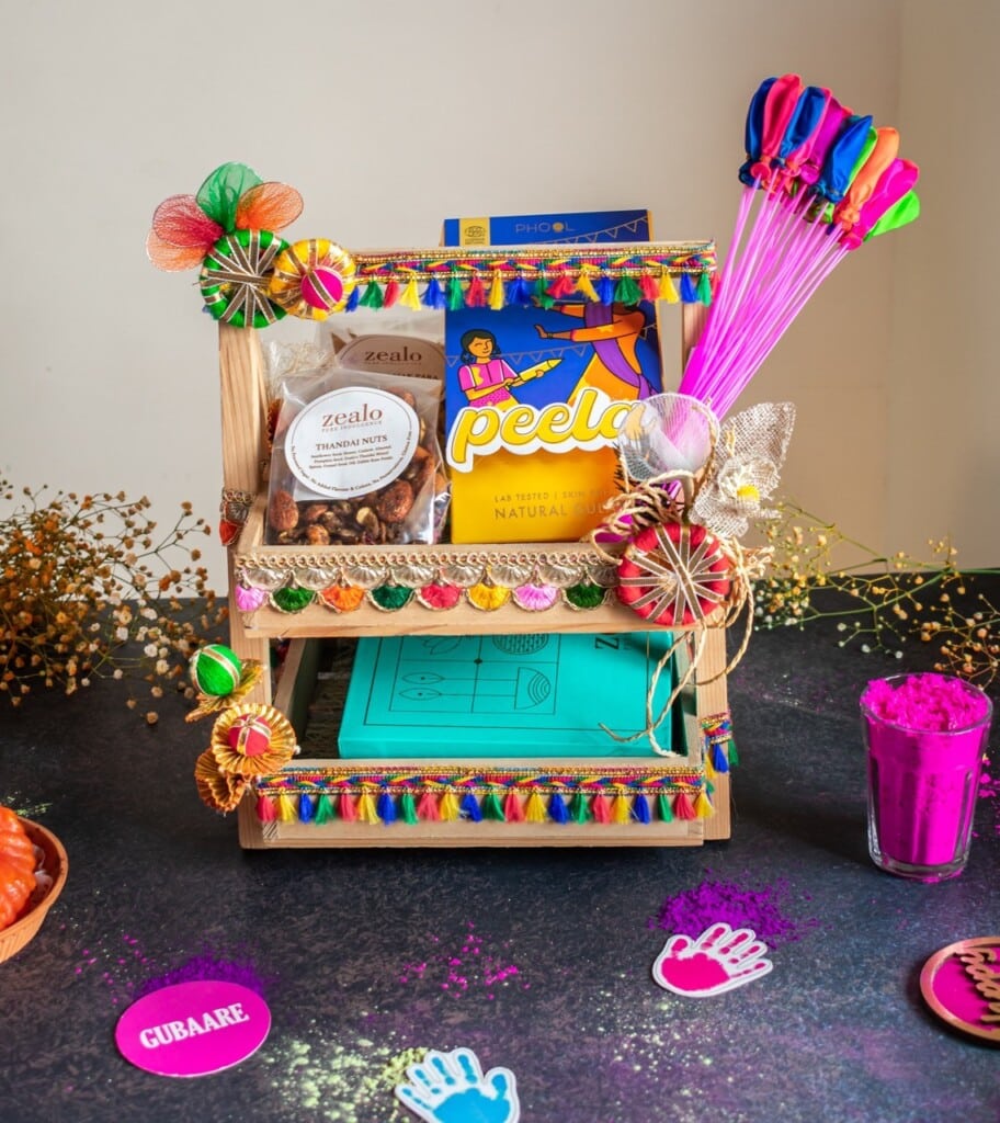 Floral Art presents A festive display features colorful balloons, a Peela snack pack, dried nuts, vibrant powders, and decorative elements on a wooden stand with a dark table background.
