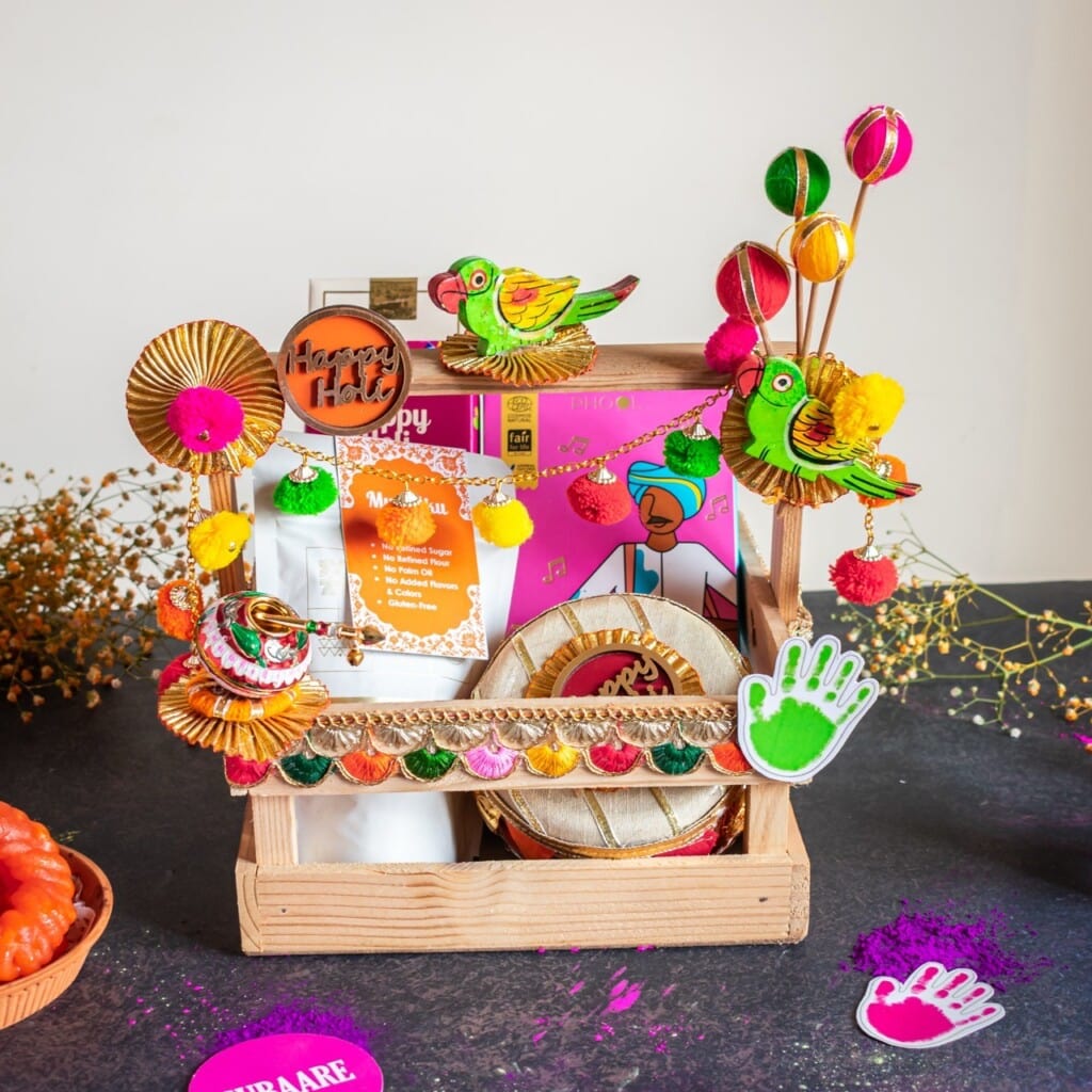 Floral Art presents A wooden basket with colorful paper birds, pom poms, handprint cutouts, a plate, flowers, and packets arranged for a festive decoration.