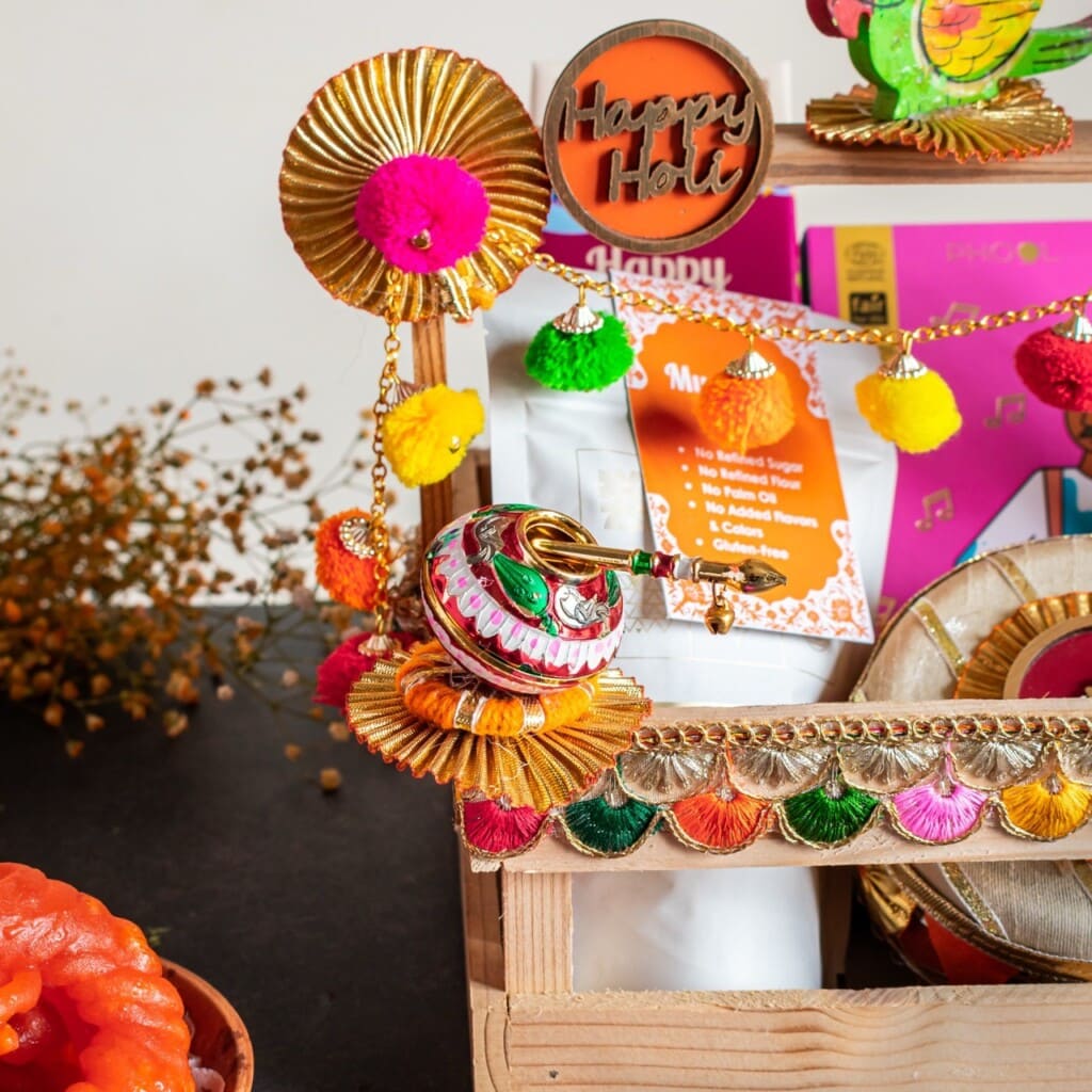 Floral Art presents Colorful festive decorations with "Happy Holi" sign, vibrant pom poms, and shimmering gold accents displayed on a wooden shelf.