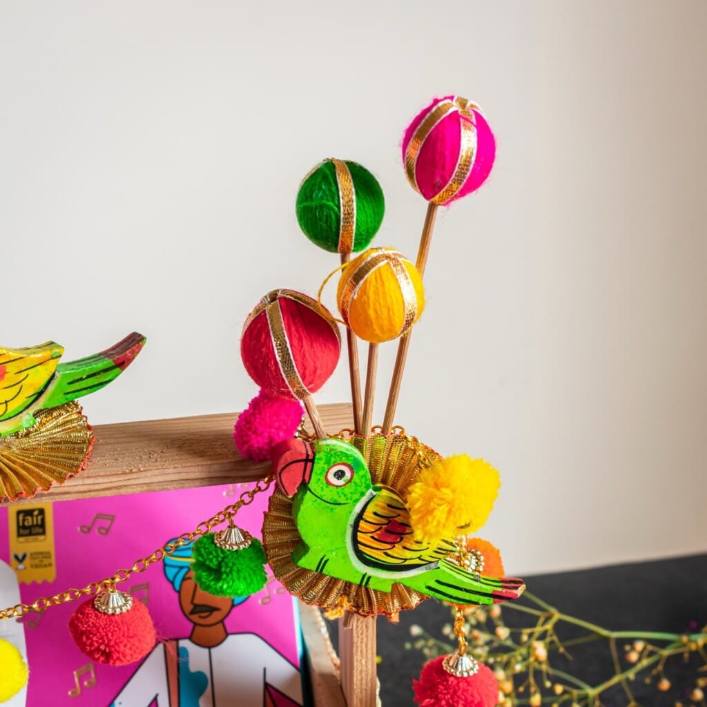 Floral Art presents Colorful Indian garland with green parrot decoration, vibrant pom poms, and tassels against a neutral background.