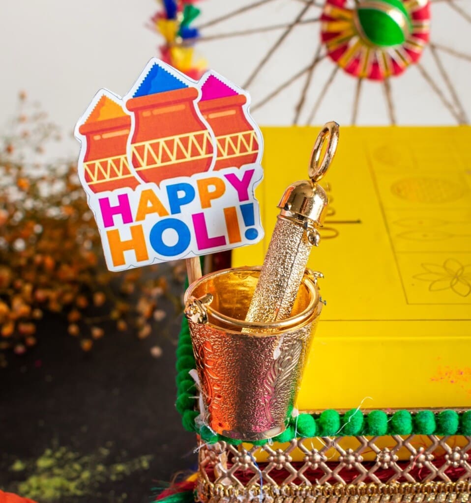 Floral Art presents Colorful "Happy Holi" sign next to a gold container with a decorative item inside, set on a yellow block with festive decorations.