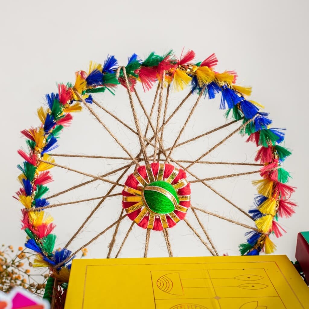 Floral Art presents Colorful, circular craft with a multicolored yarn and straw design, featuring red, yellow, blue, and green tassels and a geometric center, placed against a white background.