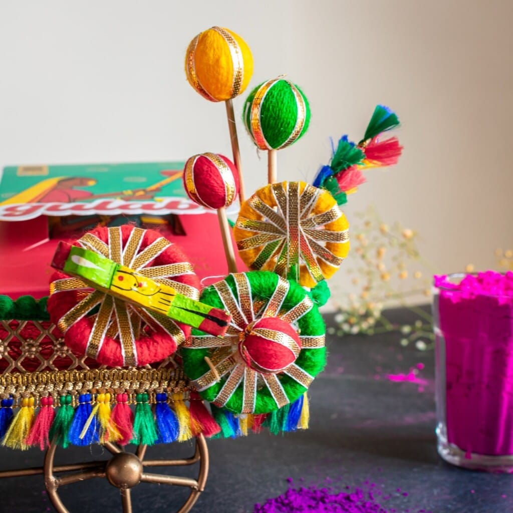 Floral Art presents Colorful decorative items with thread and cloth are on a table. Nearby, a glass holds magenta powder, and a colorful book is partially visible in the background.