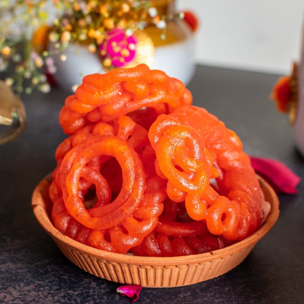 Floral Art presents A basket of orange jalebis placed on a dark surface, with decorative elements and a white pot in the background.