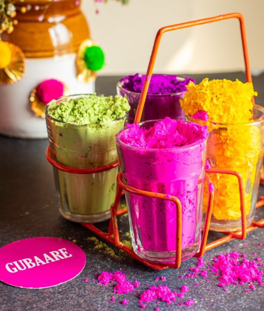 Floral Art presents Four glasses filled with bright colored powders, displayed in an orange metal holder. A round Gubaare label and decorative flowers are in the background.