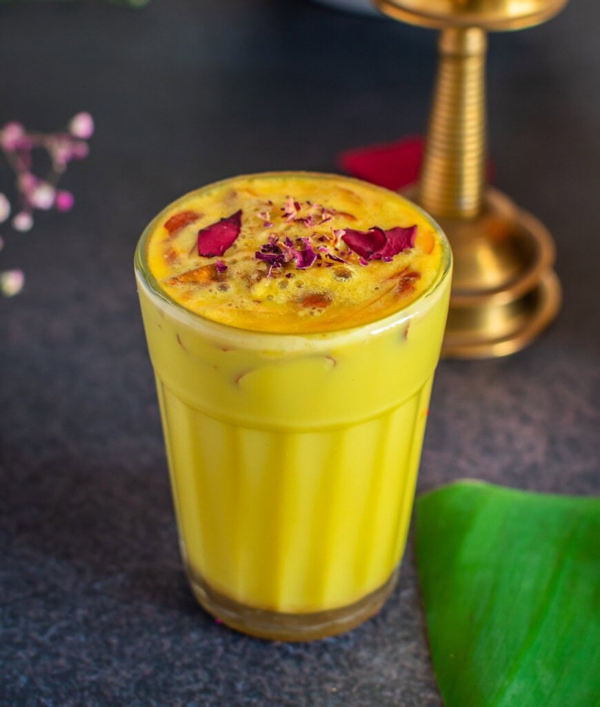 Floral Art presents A glass of yellow lassi topped with rose petals, placed on a dark surface. A brass lamp is partially visible in the background.