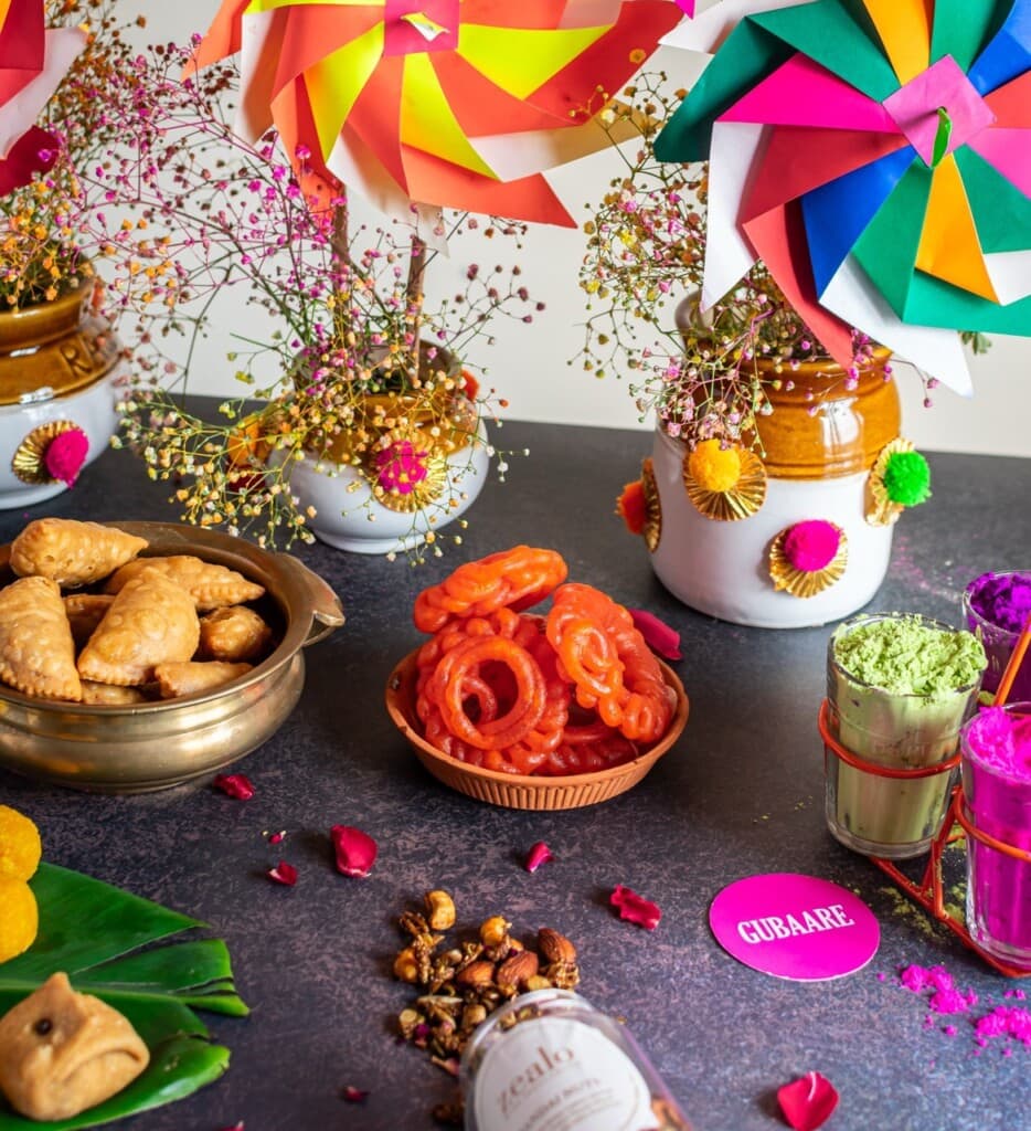 Floral Art presents Colorful pinwheels, snacks, and Holi powders are arranged on a surface. Decorated jars with flowers are also present, creating a festive atmosphere.