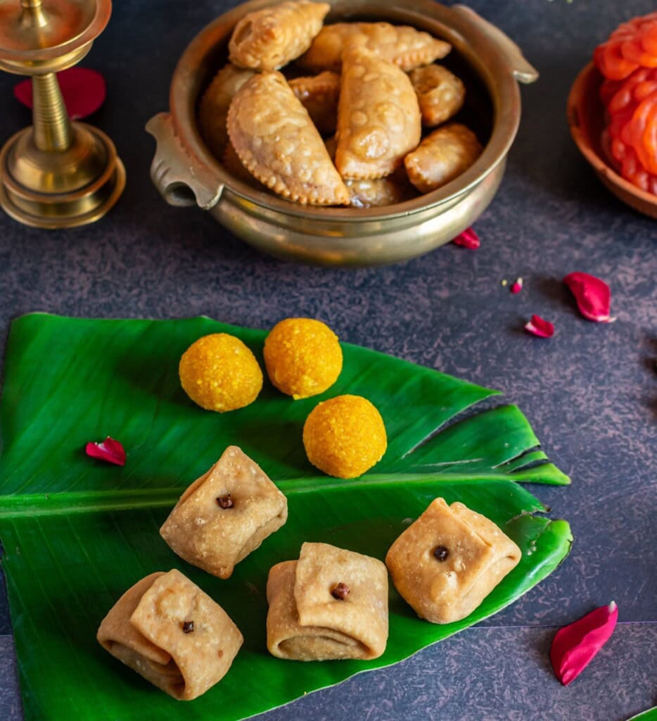Floral Art presents Assorted Indian sweets and snacks, including gujiya and laddu, are arranged on a banana leaf and brass plate, surrounded by rose petals and a brass candle holder.