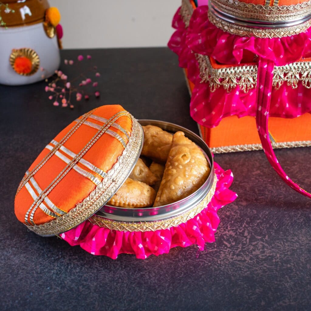 Floral Art presents Decorative tin with festive lid containing fried pastries, possibly samosas, on a dark surface. Surrounding are ribboned gift boxes in vibrant pink and orange hues.