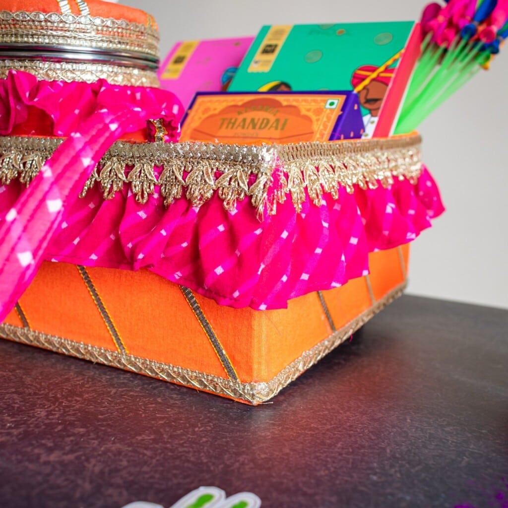 Floral Art presents Decorative orange gift box with pink fabric and ornate trim, containing colorful notebooks and packaged food items, situated on a dark surface.