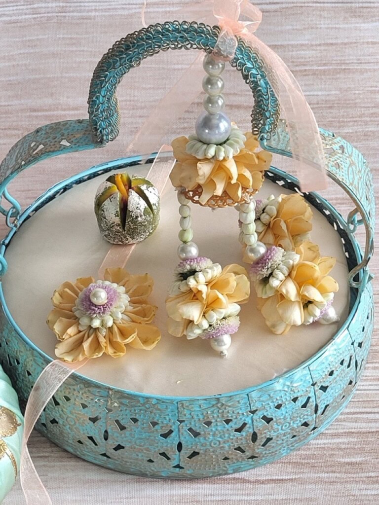 Floral Art presents Blue metal basket with a decorative handle, containing yellow artificial flower jewelry and a small green and gold ornamental piece, placed on a light colored surface.