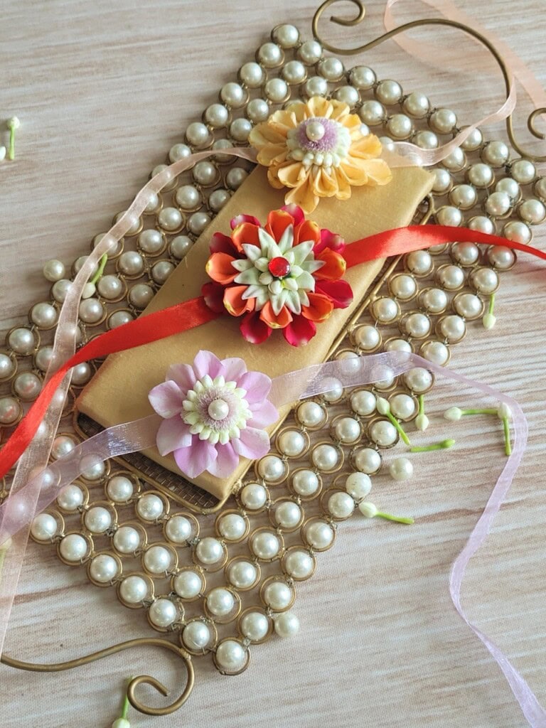 Floral Art presents A gold envelope decorated with three colorful artificial flowers and a red ribbon, placed on a beaded pearl mat with pink ribbon accents on a wooden surface.