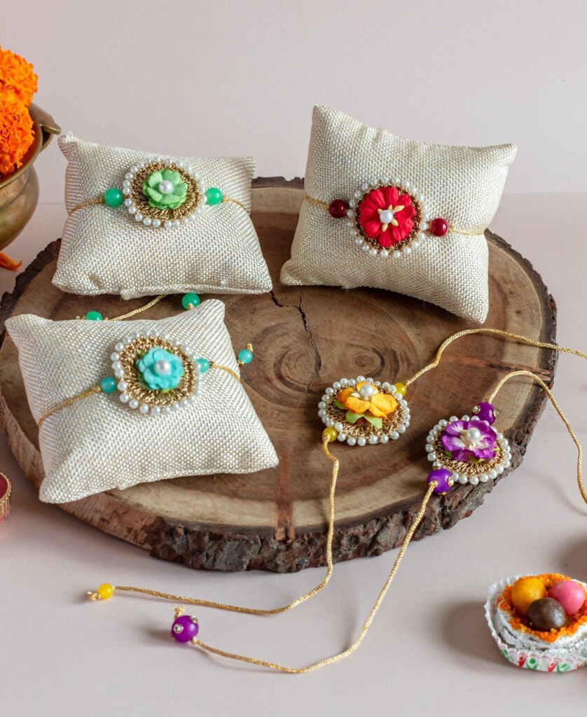 Floral Art presents Four decorative rakhis with colorful beads and floral designs are displayed on beige cushions on a wooden slab, with a bowl of marigold flowers and a small dish of sweets nearby.