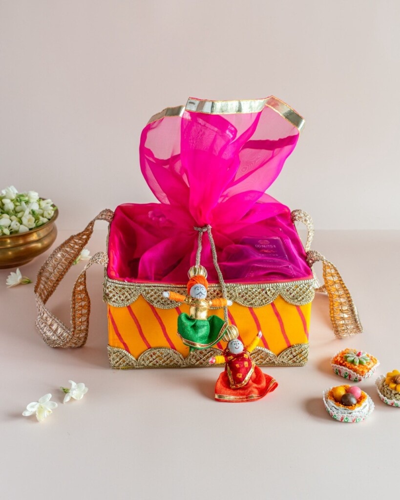Floral Art presents A decorated gift box with gold straps, bright orange and pink wrapping, adorned with two traditional doll ornaments, surrounded by small bowls of sweets and flowers.