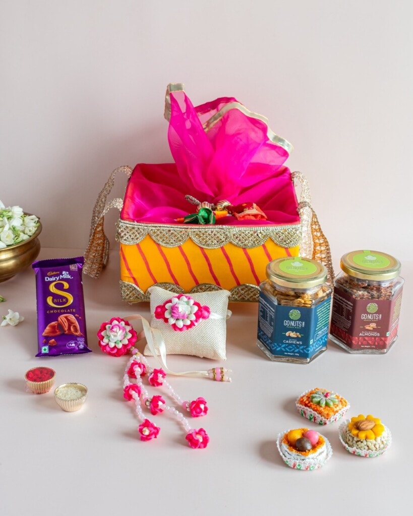 Floral Art presents A festive gift basket with sweets, jars of cookies, chocolates, floral jewelry, a small pouch, and colorful decorative items arranged on a white surface.