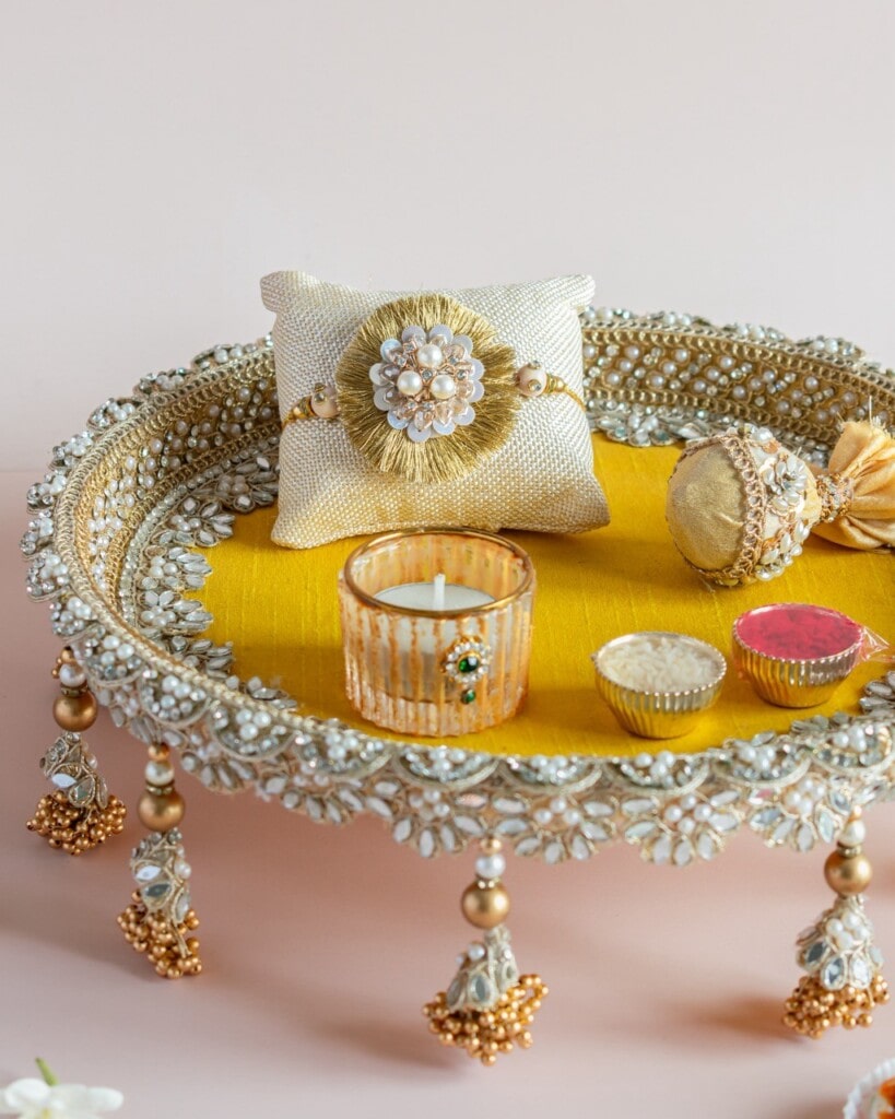Floral Art presents A decorative tray with gold and pearl accents holds a rakhi on a cushion, a candle, sweets, and small ornamental containers on a yellow cloth.