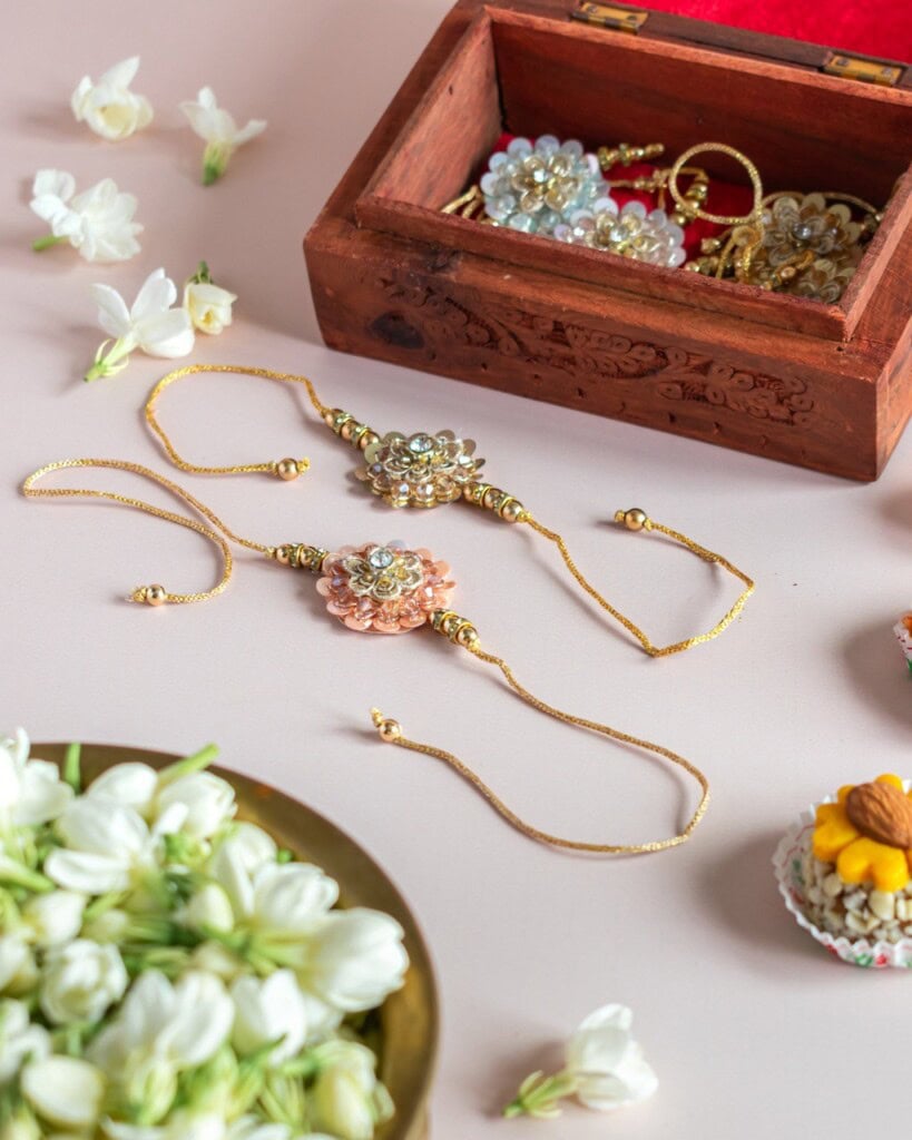 Floral Art presents Three decorative gold bracelets are displayed on a light surface next to a wooden jewelry box, white flowers, and small bowls of nuts and flower petals.