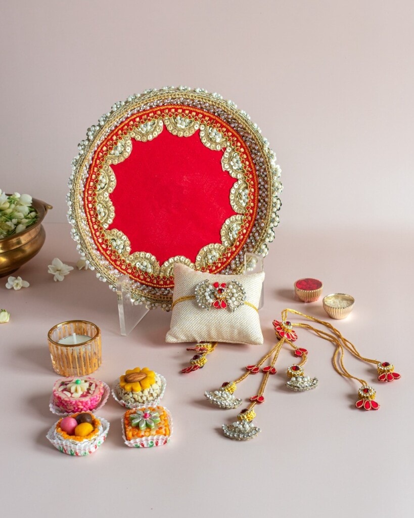 Floral Art presents A decorated red and gold thali with traditional sweets, a candle, a pillow with jewelry, and gold rakhi threads arranged on a light pink surface.