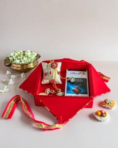Floral Art presents A red gift box contains a rakhi, a pack of Lindt chocolate, and traditional Indian sweets, with a bowl of jasmine flowers placed nearby.